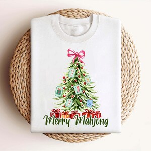 Pode incluir: Camisola branca de gola redonda com um desenho de &aacute;rvore de Natal com la&ccedil;o rosa, enfeites e caixas de presente. A &aacute;rvore &eacute; decorada com pe&ccedil;as de Mahjong. As palavras "Merry Mahjong" est&atilde;o escritas em verde abaixo dos presentes.