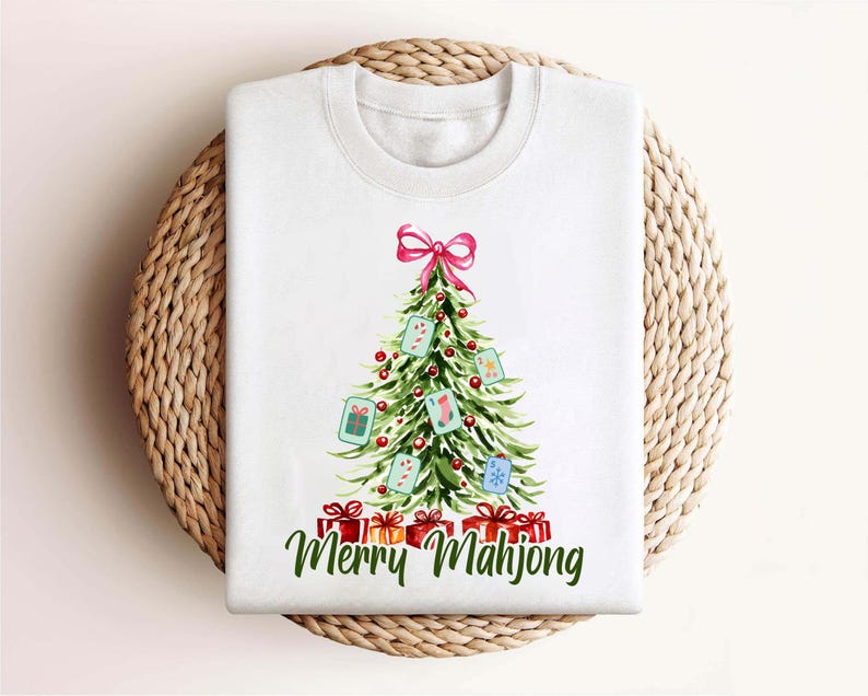 Pode incluir: Camisola branca de gola redonda com um desenho de &aacute;rvore de Natal com la&ccedil;o rosa, enfeites e caixas de presente. A &aacute;rvore &eacute; decorada com pe&ccedil;as de Mahjong. As palavras "Merry Mahjong" est&atilde;o escritas em verde abaixo dos presentes.