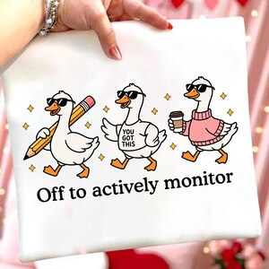 Può includere: Borsa tote bianca con tre oche dei cartoni animati con occhiali da sole. Un'oca tiene una matita, un'altra indossa una maglietta con la scritta "You Got This", e la terza ha una tazza di caffè. Il testo in basso recita "Off to actively monitor."