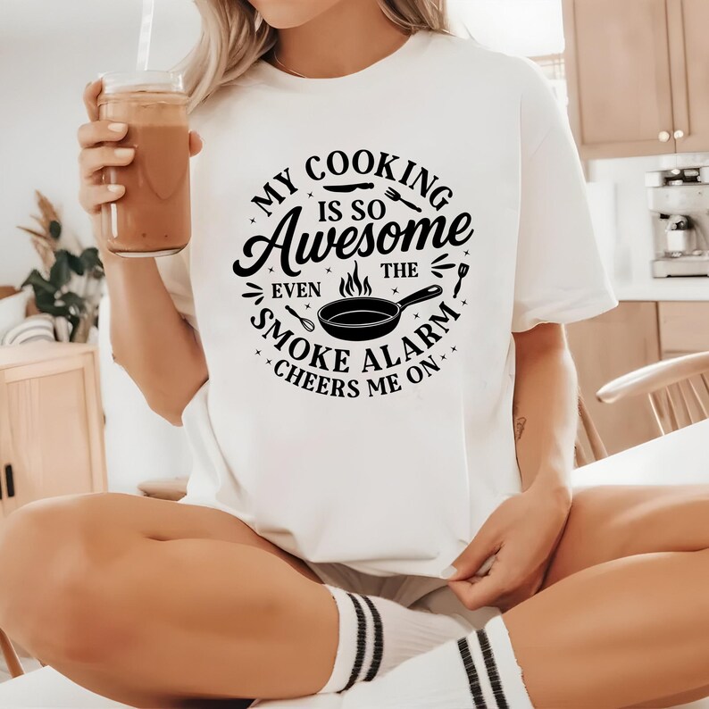 Puede incluir: Camiseta blanca con un gr&aacute;fico negro que dice "My Cooking Is So Awesome Even The Smoke Alarm Cheers Me On." El dise&ntilde;o incluye una sart&eacute;n, utensilios y llamas. La persona sostiene un vaso de l&iacute;quido marr&oacute;n.