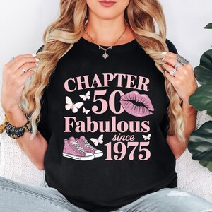 May include: Black t-shirt with pink text and graphics. The text reads "CHAPTER 50 Fabulous since 1975" with butterfly and sneaker graphics. The number 50 is in a large pink font, and a pair of pink sneakers are at the bottom.