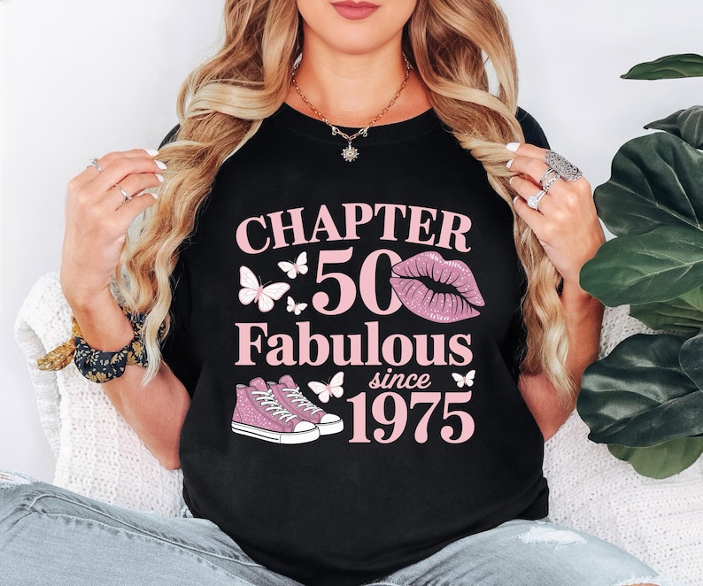 May include: Black t-shirt with pink text and graphics. The text reads "CHAPTER 50 Fabulous since 1975" with butterfly and sneaker graphics. The number 50 is in a large pink font, and a pair of pink sneakers are at the bottom.