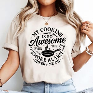 Puede incluir: Camiseta beige con un gr&aacute;fico negro que dice "My Cooking Is So Awesome Even The Smoke Alarm Cheers Me On." El dise&ntilde;o incluye una sart&eacute;n, utensilios y estrellas. La camiseta tiene mangas cortas.