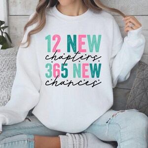 Può includere: Felpa bianca con la scritta "12 NEW chapters 365 NEW chances" in verde acqua e rosa. Il testo è in un carattere giocoso con accenti di stelle. La felpa ha una vestibilità ampia.