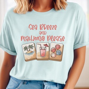 Może przedstawiać: Jasnoniebieska koszulka z r&oacute;żowym napisem "Sea Breeze and Mahjong Please". Koszulka zawiera trzy ilustrowane kafelki: tropikalną wyspę, koktajl i muszle. Projekt znajduje się z przodu koszulki.