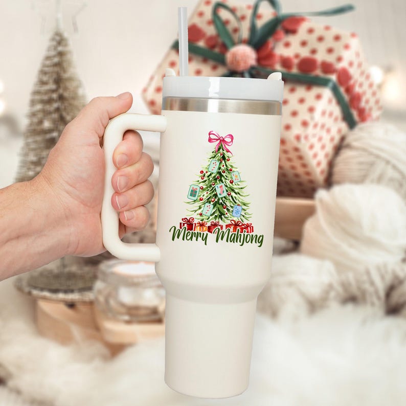Pode incluir: Um copo t&eacute;rmico branco com uma al&ccedil;a e uma palhinha. O copo apresenta um desenho de &aacute;rvore de Natal com um la&ccedil;o rosa, presentes e o texto "Merry Mahjong" em verde. O copo &eacute; segurado por uma m&atilde;o.