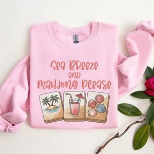 Może przedstawiać: Jasnor&oacute;żowa bluza z napisem "Sea Breeze and Mahjong Please" w koralowych literach. Poniżej znajdują się trzy ilustracje: tropikalna wyspa, koktajl i muszle. Bluza ma długie rękawy.