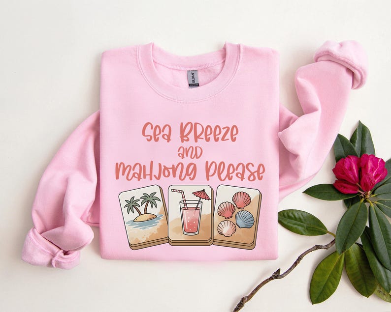 Może przedstawiać: Jasnor&oacute;żowa bluza z napisem "Sea Breeze and Mahjong Please" w koralowych literach. Poniżej znajdują się trzy ilustracje: tropikalna wyspa, koktajl i muszle. Bluza ma długie rękawy.