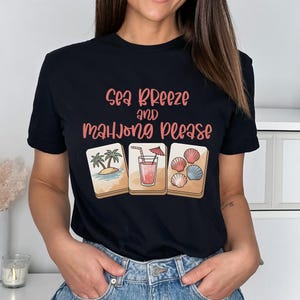 Może przedstawiać: Czarny t-shirt z napisem "Sea Breeze and Mahjong Please" w r&oacute;żowych i brzoskwiniowych literach. Koszulka przedstawia trzy ilustrowane kafelki do mahjonga: scenę plażową, koktajl i muszle. Modelka ma na sobie niebieskie dżinsy.