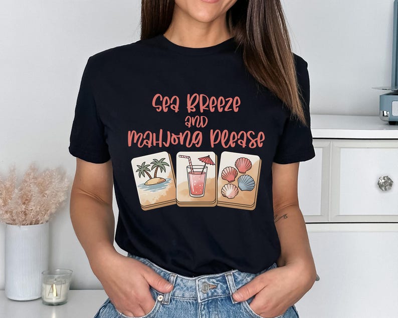 Może przedstawiać: Czarny t-shirt z napisem "Sea Breeze and Mahjong Please" w r&oacute;żowych i brzoskwiniowych literach. Koszulka przedstawia trzy ilustrowane kafelki do mahjonga: scenę plażową, koktajl i muszle. Modelka ma na sobie niebieskie dżinsy.