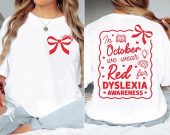 May include: A white t-shirt featuring a red bow graphic on the front left chest with the text Dyslexia Awareness. The back displays a large red design with a bow, a book, a lightbulb, a brain, and the text In October we wear Red for DYSLEXIA AWARENESS.