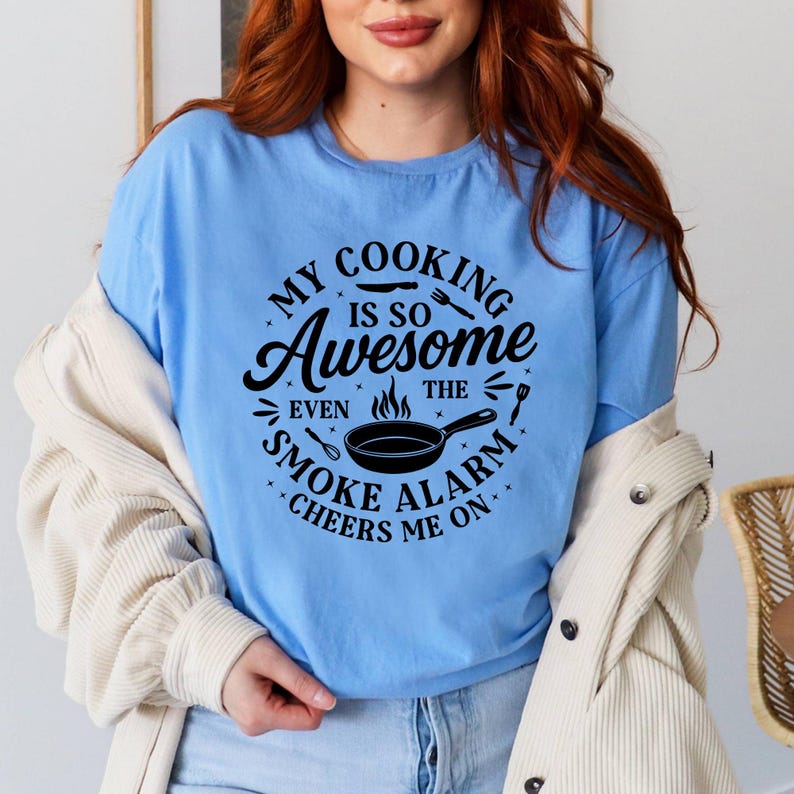 Puede incluir: Camiseta azul claro con el texto "MY COOKING IS SO Awesome EVEN THE SMOKE ALARM CHEERS ME ON." El dise&ntilde;o incluye una sart&eacute;n y utensilios. La camiseta es de tejido suave.