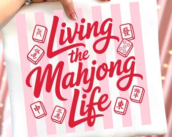 May include: A white square pillow with pink vertical stripes and the text "Living the Mahjong Life" in red script. The design includes red Mahjong tiles. The pillow is held by a person.