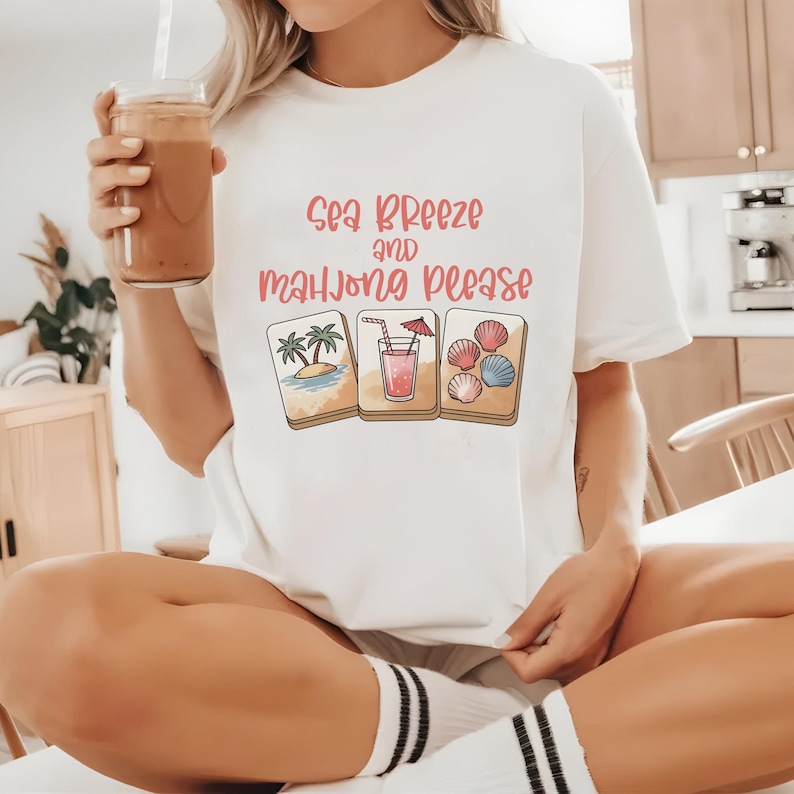 Może przedstawiać: Biała koszulka z r&oacute;żowym napisem "Sea Breeze and Mahjong Please". Koszulka przedstawia trzy ilustrowane kafelki: scenę z wyspą, koktajl i muszle. Osoba trzyma szklankę brązowego napoju.