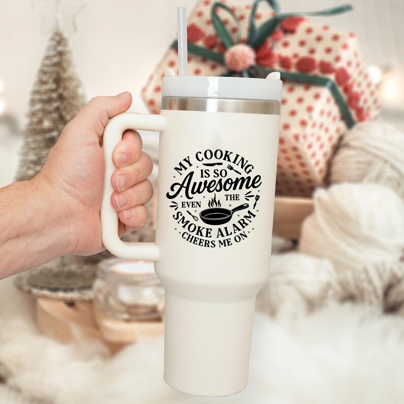 Puede incluir: Un vaso t&eacute;rmico blanco con asa y pajita. El vaso tiene una tapa blanca y presenta el texto negro "MY COOKING IS SO Awesome EVEN THE SMOKE ALARM CHEERS ME ON.". El vaso est&aacute; sujeto por una mano.