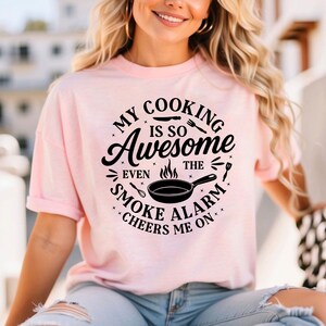 Puede incluir: Camiseta rosa claro con el texto "MY COOKING IS SO Awesome EVEN THE SMOKE ALARM CHEERS ME ON". El dise&ntilde;o incluye una sart&eacute;n, cubiertos y estrellas. La camiseta est&aacute; siendo usada.