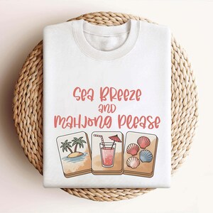 Może przedstawiać: Biała bluza z okrągłym dekoltem z napisem "Sea Breeze and Mahjong Please" w kolorze koralowym. Projekt zawiera ilustracje tropikalnej wyspy, koktajlu i muszli. Bluza jest prezentowana na tkanej, okrągłej macie.