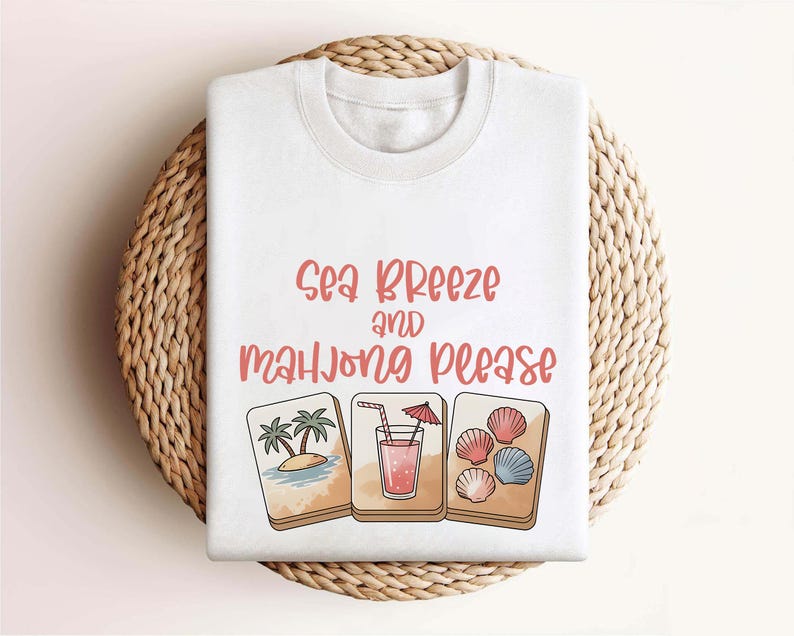 Może przedstawiać: Biała bluza z okrągłym dekoltem z napisem "Sea Breeze and Mahjong Please" w kolorze koralowym. Projekt zawiera ilustracje tropikalnej wyspy, koktajlu i muszli. Bluza jest prezentowana na tkanej, okrągłej macie.