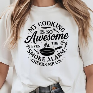 Puede incluir: Camiseta blanca con un dise&ntilde;o negro que dice: "MY COOKING IS SO Awesome EVEN THE SMOKE ALARM CHEERS ME ON." El dise&ntilde;o incluye una sart&eacute;n, llamas y utensilios de cocina.