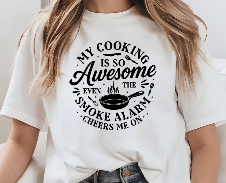 Puede incluir: Camiseta blanca con un dise&ntilde;o negro que dice: "MY COOKING IS SO Awesome EVEN THE SMOKE ALARM CHEERS ME ON." El dise&ntilde;o incluye una sart&eacute;n, llamas y utensilios de cocina.
