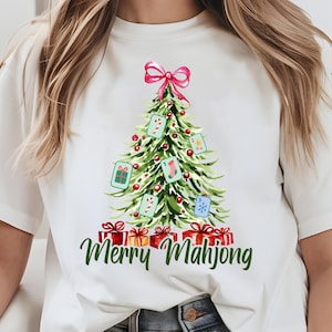 Pode incluir: T-shirt branca com um desenho de &aacute;rvore de Natal em aquarela. A &aacute;rvore &eacute; decorada com pe&ccedil;as de Mahjong, enfeites vermelhos e um la&ccedil;o rosa. As palavras "Merry Mahjong" est&atilde;o escritas em verde por baixo.
