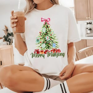 Pode incluir: Camiseta branca com um desenho de &aacute;rvore de Natal em aquarela, decorada com pe&ccedil;as de Mahjong e um la&ccedil;o rosa. A frase "Merry Mahjong" est&aacute; escrita em verde abaixo da &aacute;rvore, com caixas de presente vermelhas na base.