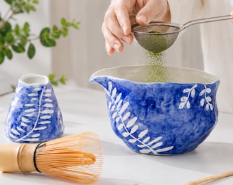 Może przedstawiać: Niebiesko-biała ceramiczna miseczka do matchy z ręcznie malowanym wzorem liści, pasujący ceramiczny dzbanek, bambusowa miotełka oraz bambusowa łyżeczka do herbaty. Metalowe sitko służy do przesiewania zielonego proszku matcha do miseczki.