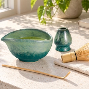 May include: A ceramic matcha bowl with a teal and green gradient glaze, a matching teal whisk holder, a bamboo whisk, and a bamboo tea scoop. The bowl features a pouring spout and contains green tea. A small beige cloth is placed under the whisk.