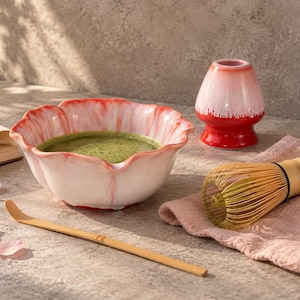 Può includere: Una ciotola per matcha in ceramica con bordo smerlato e finitura sfumata rosa e bianca contenente tè verde. Accanto si trovano un frustino in bambù, un cucchiaio in bambù e un supporto in ceramica rosa. Un piccolo libro e petali rosa sono vicini.