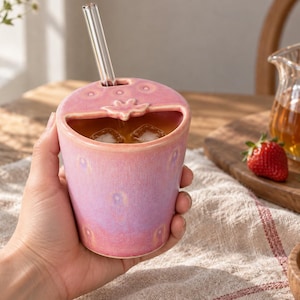May include: A pink ceramic tumbler shaped like a strawberry with a textured surface, a wide opening at the top, and a small green leaf detail. The cup includes a clear glass straw inserted through a dedicated hole in the lid, containing liquid and ice.