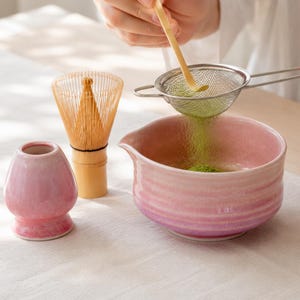 May include: A pink ceramic matcha bowl with a pouring spout, a matching pink ceramic whisk holder, and a bamboo whisk. Green matcha powder is being sifted through a fine-mesh metal strainer into the bowl using a bamboo scoop for preparation.