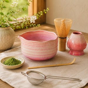 Könnte beinhalten: Ein Matcha-Teeset bestehend aus einer rosafarbenen Keramikschale mit gewelltem Rand, einem Bambusbesen, einem rosafarbenen Besenhalter, einem Bambuslöffel, einer kleinen Keramikschale mit grünem Matchapulver und einem Edelstahlsieb.