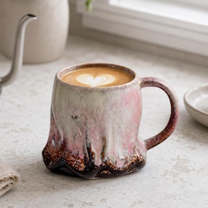 May include: A ceramic mug with a reactive glaze featuring a gradient of cream, soft pink, and deep dark brown at the base. The mug has a wide, rounded body with a curved handle and contains a coffee beverage with a heart-shaped latte art design.