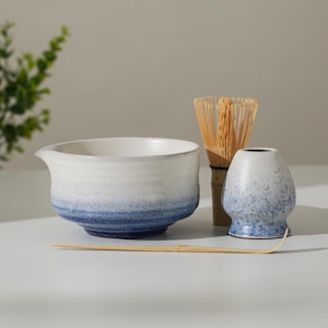May include: A matcha tea set comprising a white and blue ceramic bowl with a spout, a bamboo whisk, a small ceramic whisk holder, and a bamboo scoop. The bowl has a gradient of white to blue. The whisk is made of thin bamboo.