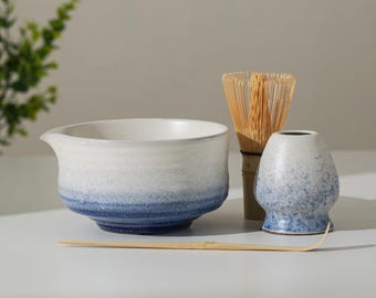 May include: A matcha tea set featuring a white and blue ceramic bowl with a spout, a bamboo whisk, a small ceramic whisk holder, and a bamboo scoop. The bowl has a gradient of white to blue. The whisk is made of thin bamboo.
