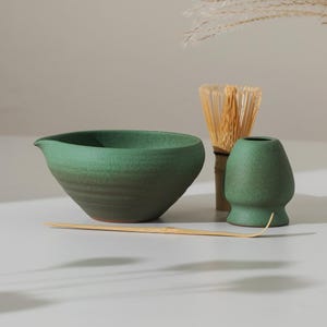 May include: A green ceramic matcha bowl with a spout, a bamboo whisk, a matcha whisk holder, and a bamboo scoop. The matcha set is arranged on a light-colored surface, with dried ornamental grass in the background.