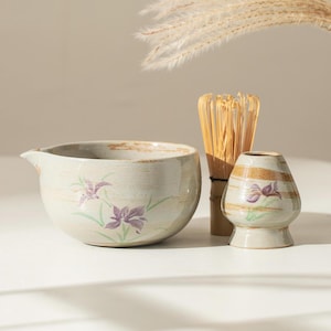 May include: A white ceramic matcha bowl with a floral design, a bamboo matcha whisk, and a white ceramic matcha cup with a floral design. The items are arranged on a white surface.