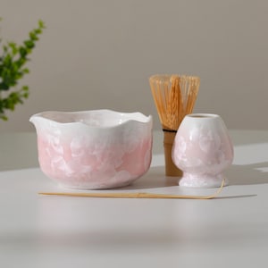 Puede incluir: Un juego de té matcha con un cuenco de cerámica rosa y blanco, un batidor, una pequeña taza de té de cerámica rosa y blanca y una cuchara de bambú. El cuenco tiene un borde ondulado y un efecto de color degradado. El batidor es de bambú fino.