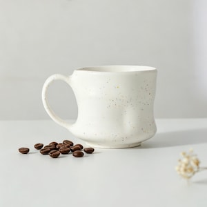 Puede incluir: Una taza de cerámica blanca con un acabado moteado que presenta pequeños puntos de colores. La taza tiene un asa redondeada y una forma orgánica ligeramente irregular. Un pequeño montón de granos de café tostados se sitúa a la izquierda.