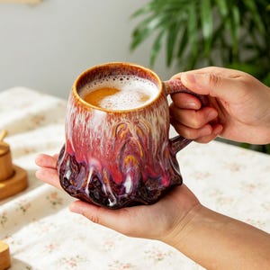 May include: A ceramic mug with a unique, textured design in shades of red, brown, and white. The mug is filled with a frothy beverage. The handle is integrated into the design. The mug is held in a person's hands.