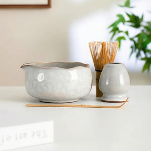 Puede incluir: Un cuenco de matcha de cerámica blanca con borde ondulado, un batidor de bambú, un pequeño soporte de matcha de cerámica y una cuchara de bambú están dispuestos sobre una superficie blanca. Los artículos son para preparar té matcha.