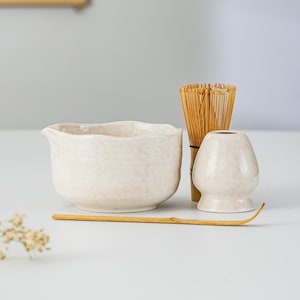 Può includere: Una ciotola per matcha in ceramica con smalto maculato rosa chiaro e beccuccio, accompagnata da un frustino in bambù, un supporto in ceramica abbinato per il frustino e un lungo e sottile cucchiaio da tè in bambù su una superficie piana.
