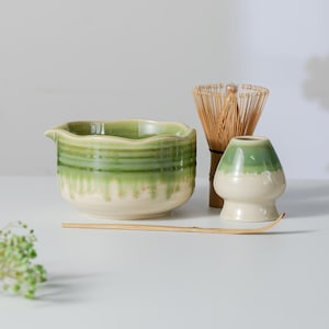 Könnte beinhalten: Eine Matcha-Schale aus Keramik mit grün-cremefarbener Tropfglasur, ein Bambusbesen, ein Bambuslöffel und ein Keramikhalter für den Besen. Die Schale hat einen gewellten Rand und einen Ausguss, während der Halter die grüne und cremefarbene Glasur aufweist.