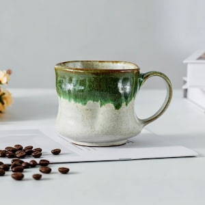 May include: A handmade ceramic mug with a green and off-white glaze. The mug has a rounded body and a simple handle. Coffee beans are scattered on the table near the mug.