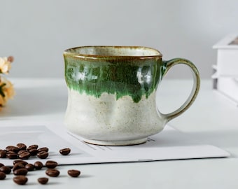 May include: A handmade ceramic mug with a green and off-white glaze. The mug has a rounded body and a simple handle. Coffee beans are scattered on the table near the mug.