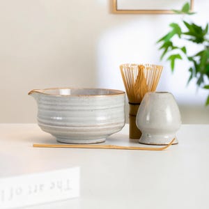 May include: A matcha tea set featuring a light gray ceramic bowl with a spout, a bamboo whisk, a whisk holder, and a bamboo scoop. The set is on a white surface, with a framed botanical print in the background.