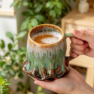 Peut inclure: Une tasse en céramique faite à la main avec un glaçage unique. La tasse présente un bord marron, un corps vert et blanc et une base marron. Elle est remplie d'un liquide brun clair et tenue par des mains. La tasse a une anse verte.