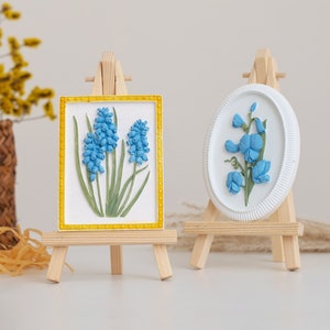 May include: Two small wooden easels, each displaying a framed floral artwork. The easel on the left features a blue flower with green leaves in a yellow frame. The easel on the right features a blue flower with green leaves in a white oval frame.