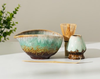 May include: A matcha tea set featuring a ceramic bowl with a spout, a bamboo whisk, a whisk holder, and a bamboo scoop. The bowl has a brown base with green and white accents. The whisk and scoop are made of bamboo.