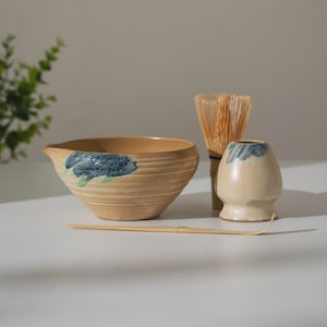 May include: A matcha tea set featuring a beige ceramic bowl, a whisk, a small cup, and a bamboo scoop. The bowl and cup have blue and green floral designs. The whisk is made of bamboo.